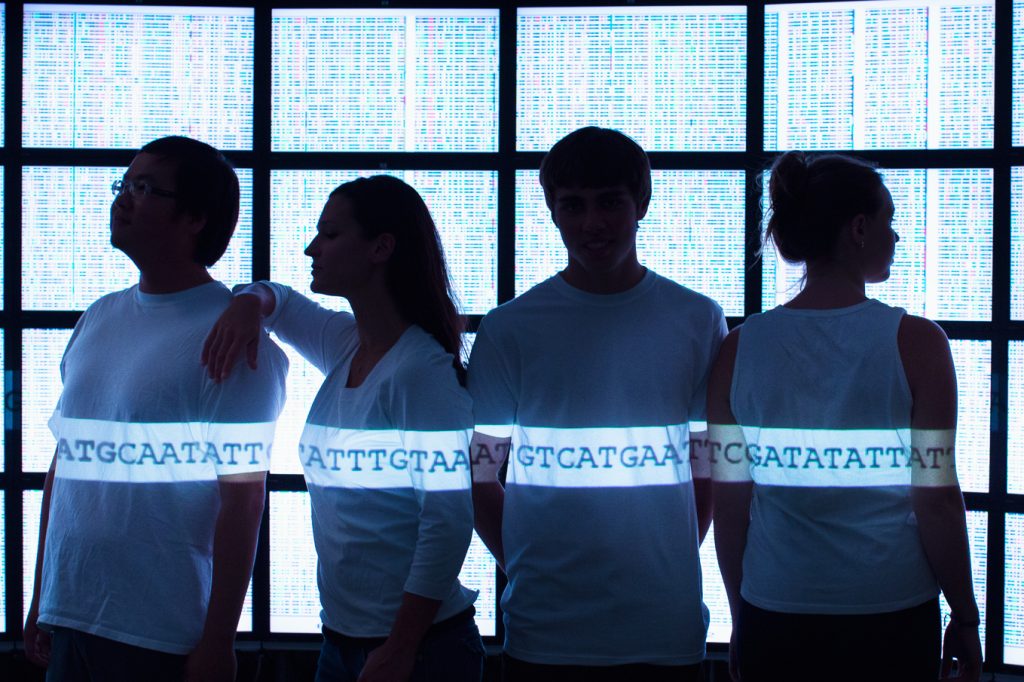 A photo of four people in white shirts standing in front of screens depicting the genetic code. An artistic representation of the Human Genome Project.