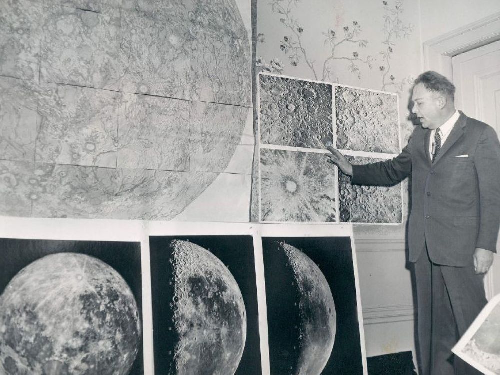 A black and white photo of a man standing in front of huge diagrams of the moon.