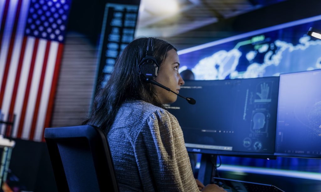 A student with a headset sits at a desk with a computer featuring a map. An American flag hangs on the wall behind her.